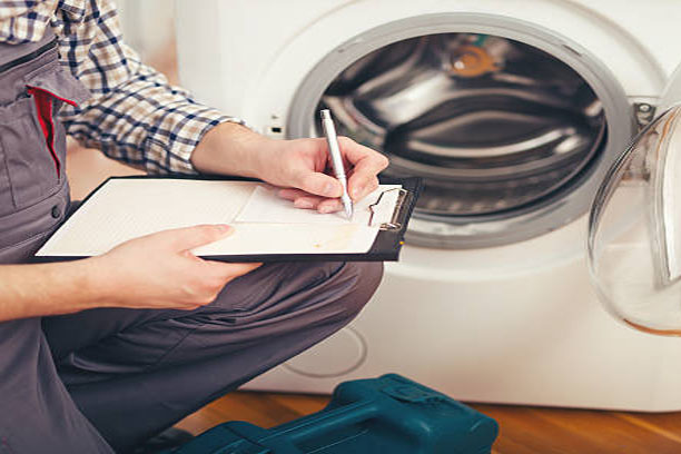 A man sits in front of a washing machine, writing notes on a clipboard with a focused expression.